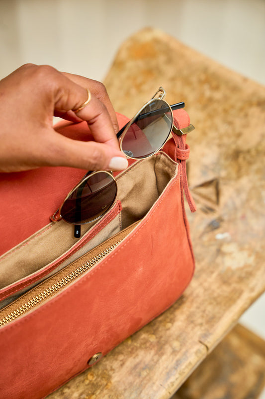 Person placing sunglasses into a red suede leather handbag on a wooden surface