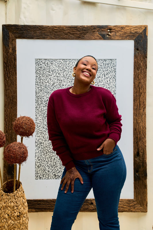 Woman in a maroon shani knit sweater and blue jeans standing in front of a framed artwork.