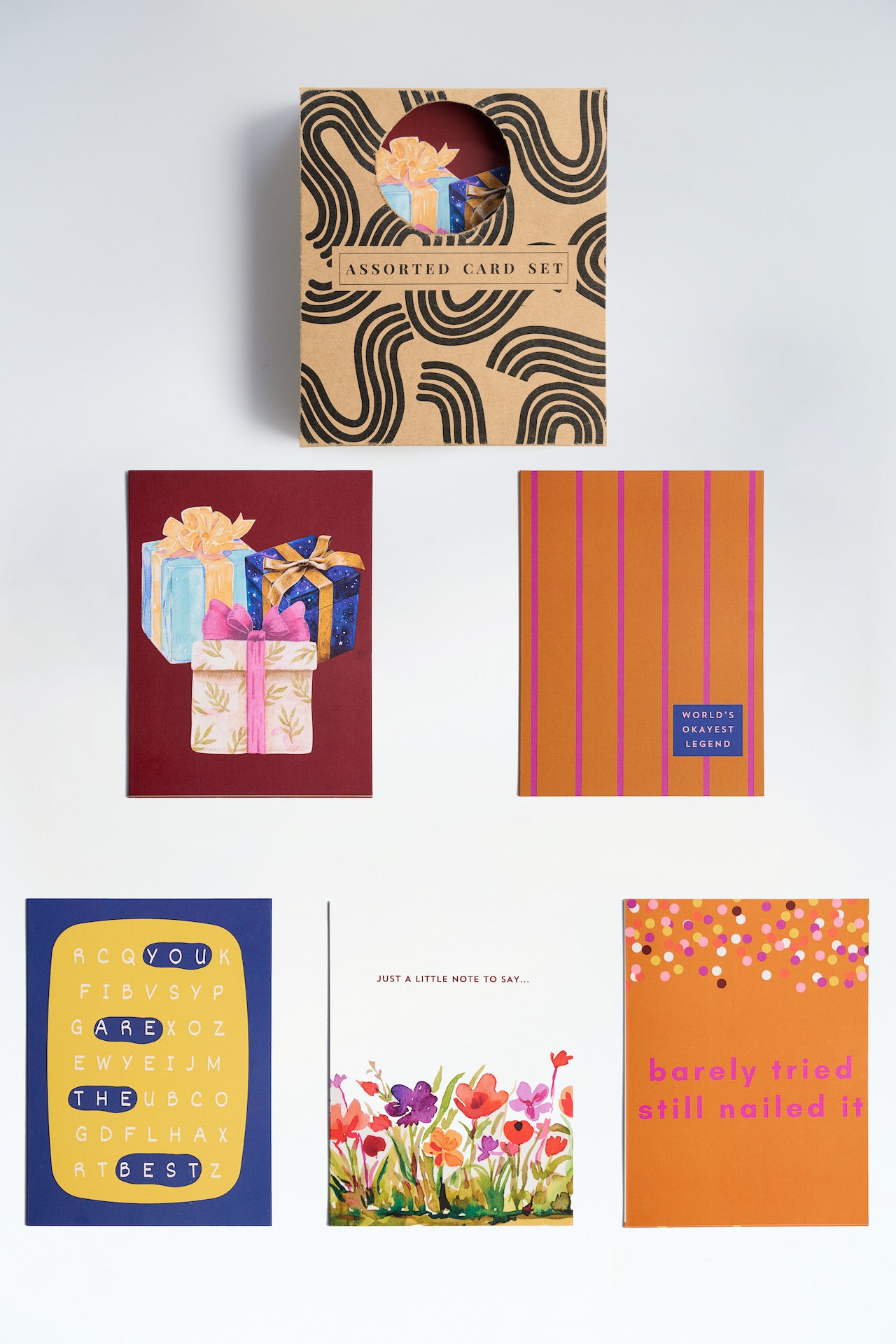 Assorted stationery gift set featuring "World's Okayest Legend" card, rainbow patterns, and retro cassette designs on a wooden table with gold gift wrap.